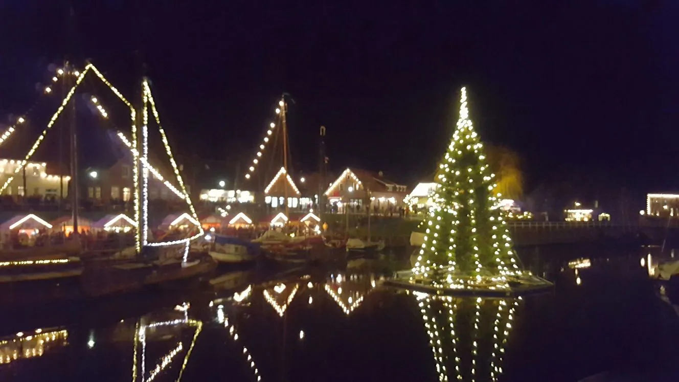 Museumshafen Carolinensiel am Abend: Plattbodenschiffe mit Lichterketten, ein beleuchteter Weihnachtsbaum auf dem Wasser und Marktbuden am Ufer