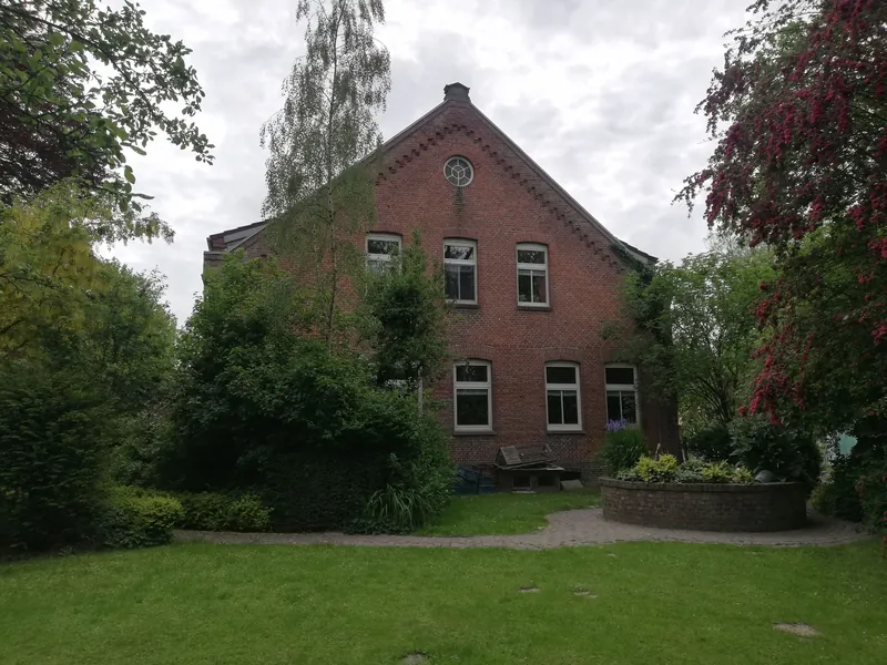 Giebel des Haupthauses mit rundem Fenster, davor der Brunnen mit Blumen und der Rasen