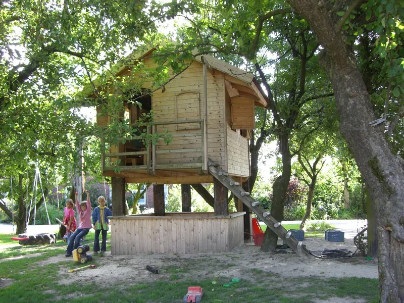 Das Stelzenhaus aus Holz mit Leiter und Sandkasten unter Apfelbäumen, daneben Kinder an der Schaukel